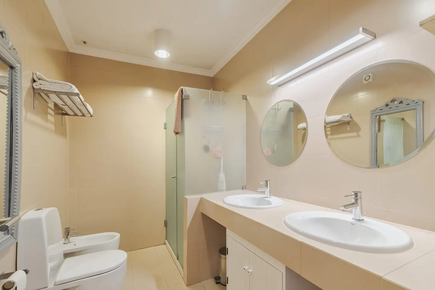 Bathroom with two sinks, round mirrors, and a glass shower. Beige walls and cabinets. Toilet and bidet on the left. Towels on a rack.