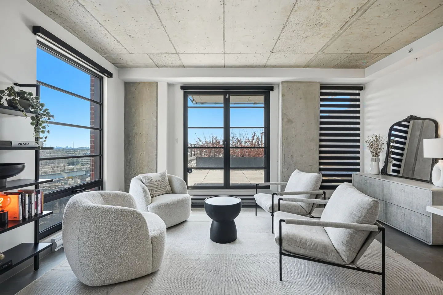 Bright living room with concrete ceiling, large windows, and four chairs. Two white, two gray. Black side table and bookshelf.