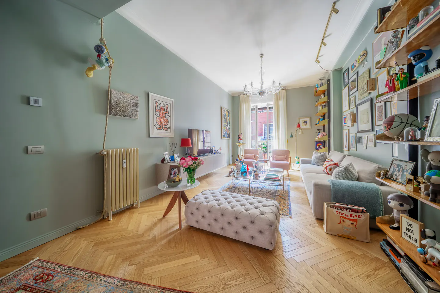 Living room with light green walls, wood floors, white sofa, tufted ottoman, and shelves filled with art and objects.