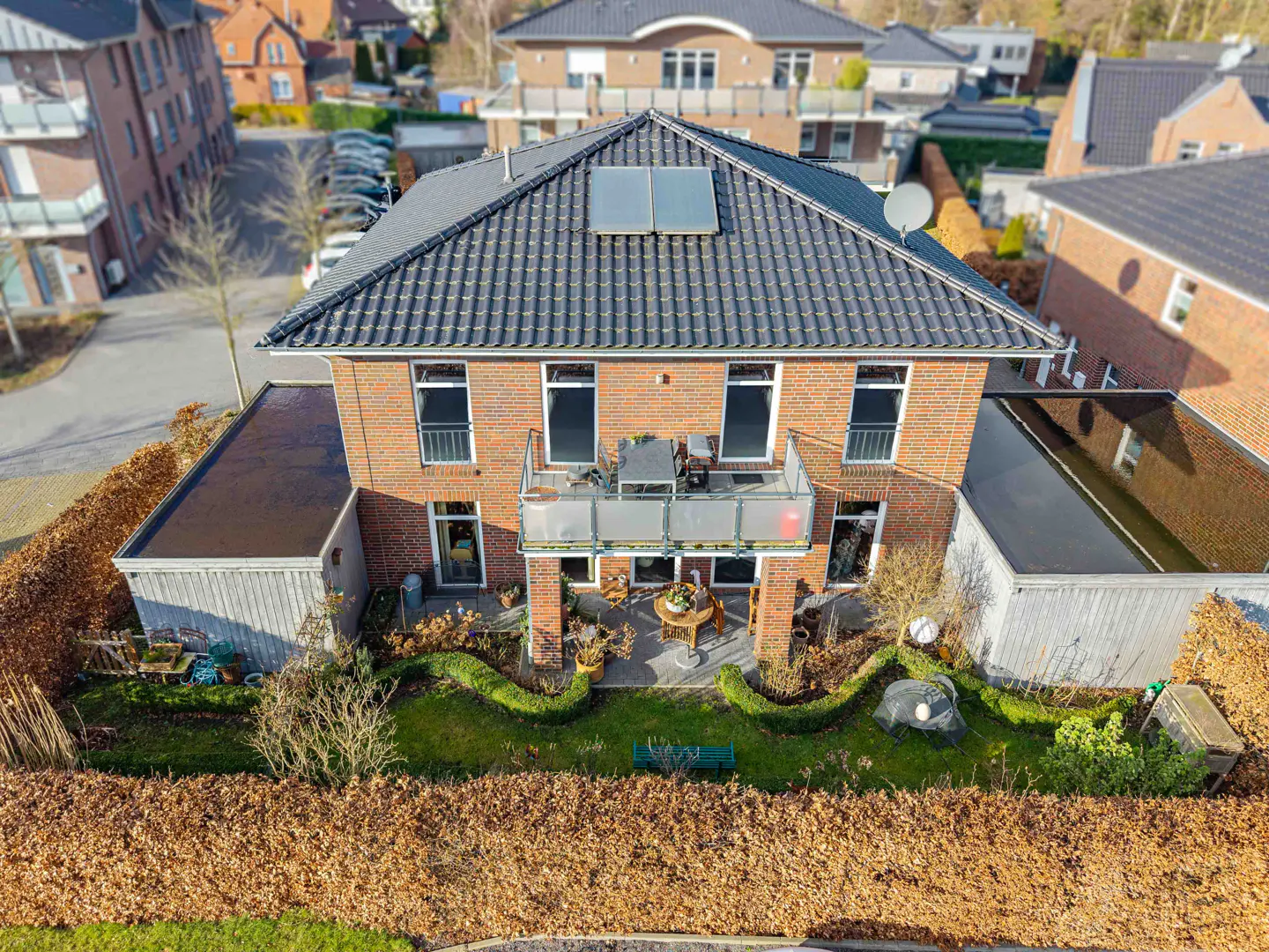 Aerial view of a two-story brick house with a balcony, dark roof, and solar panels, surrounded by a green lawn and hedges.