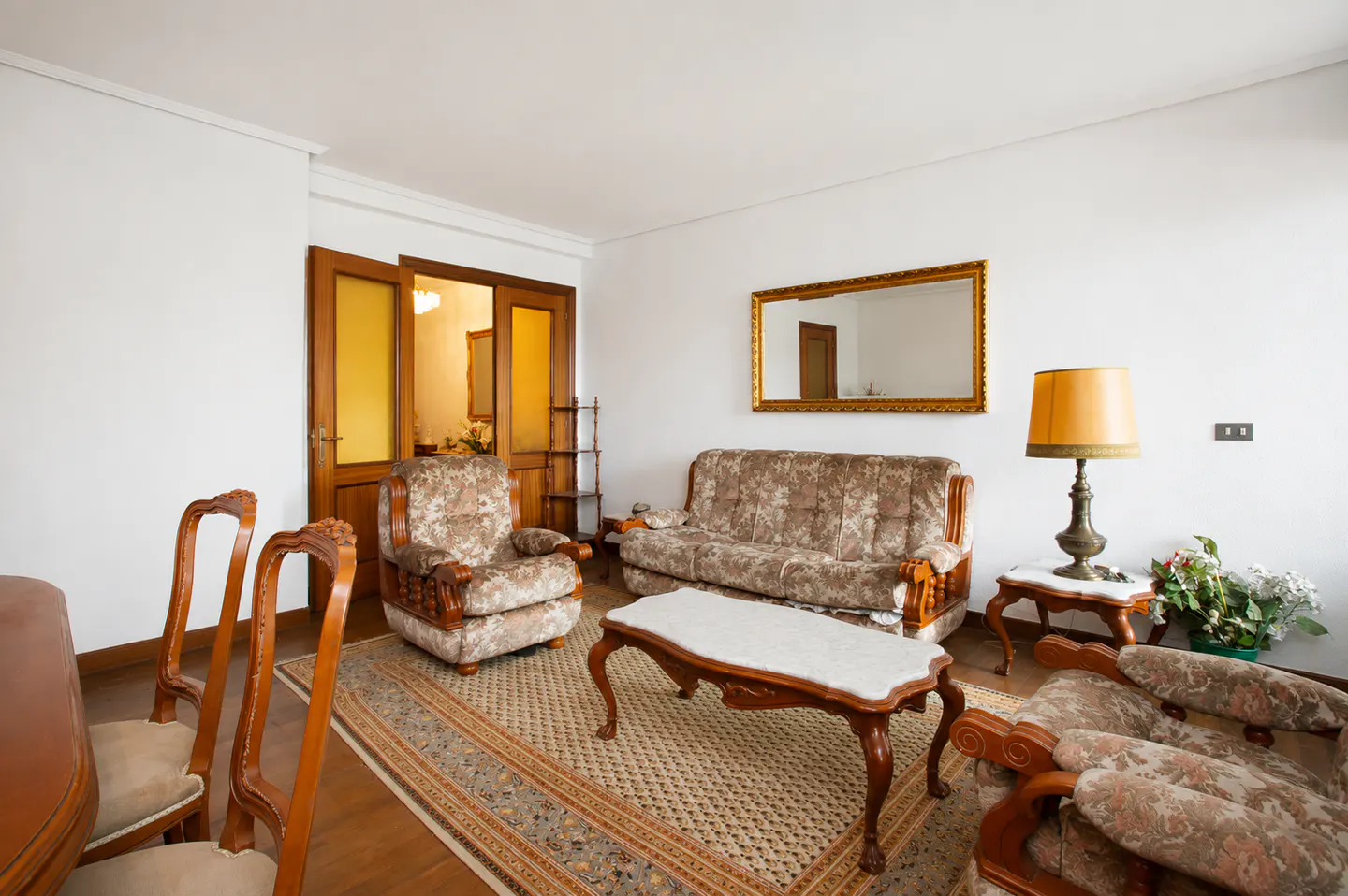 A living room with white walls, a floral sofa and armchair, a wooden coffee table, and a gold-framed mirror.