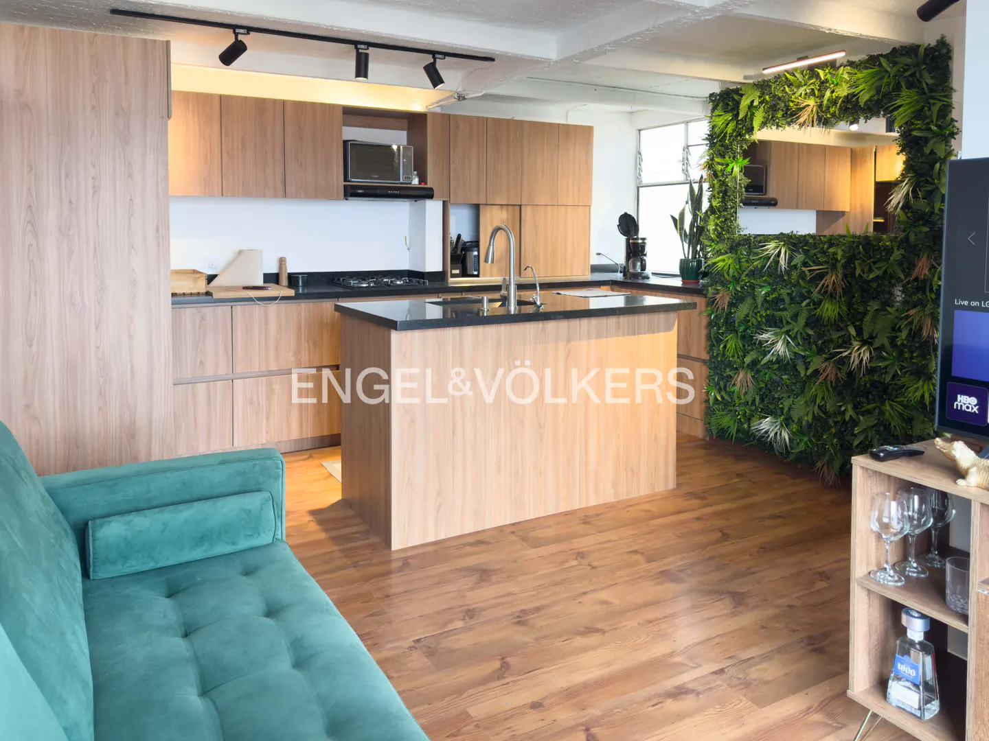 Open-concept kitchen with wood cabinets, black countertops, and a green living wall. A teal sofa sits in the foreground.