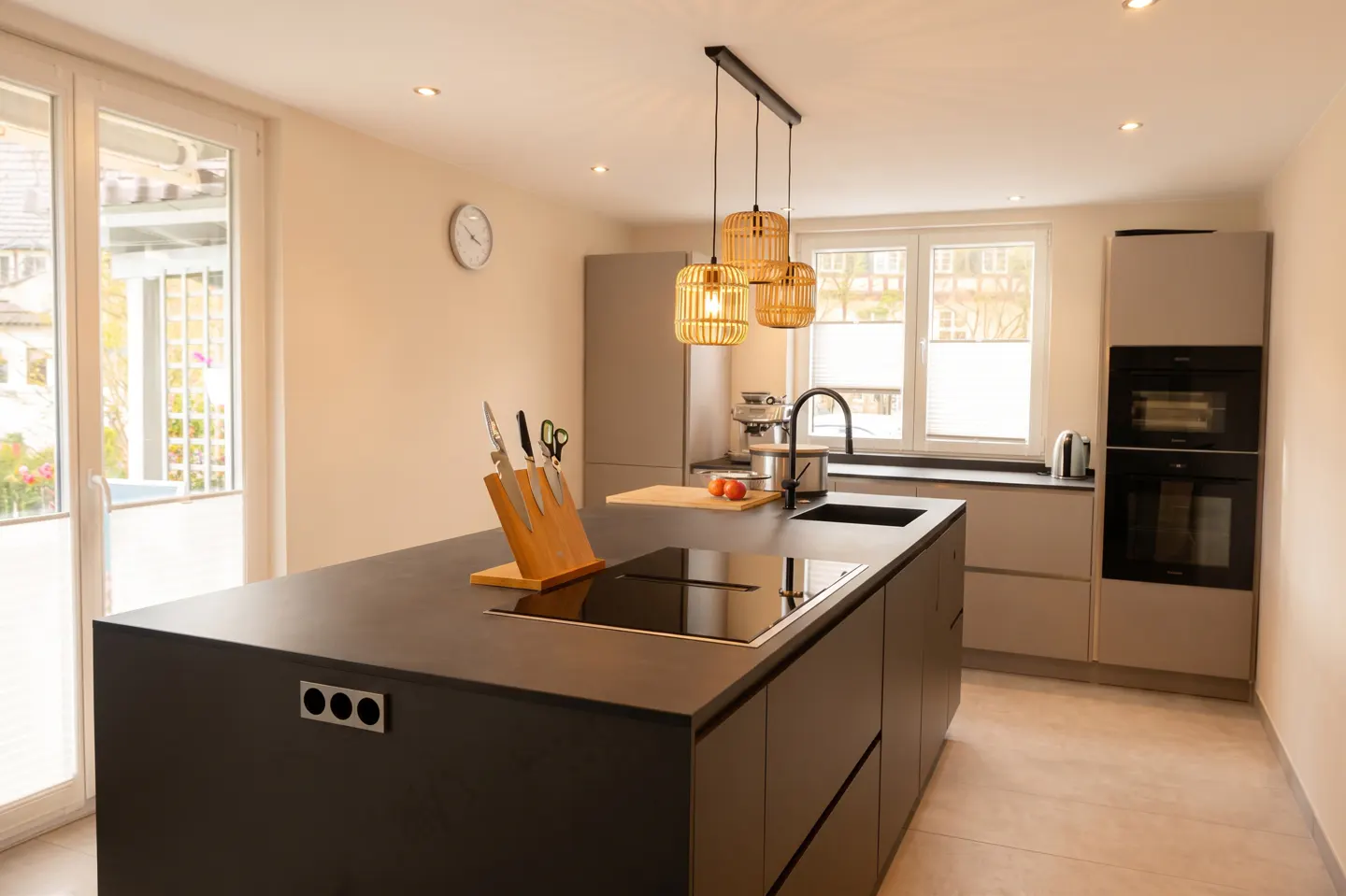 Modern kitchen with a large black island, wooden knife block, and three hanging rattan pendant lights.