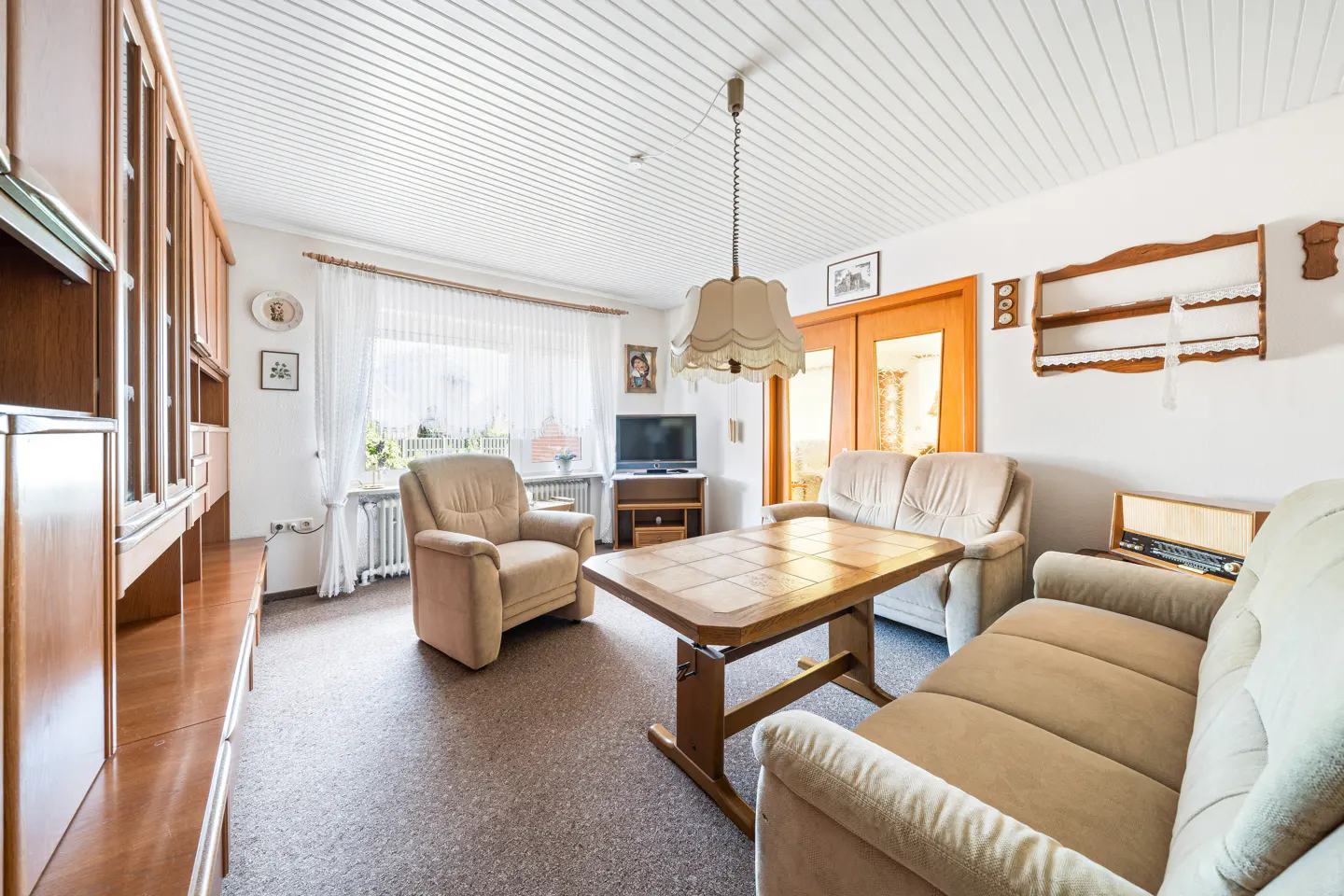 Living room with beige sofas, a wooden coffee table, a hanging lamp, and a large wooden wall unit on a carpeted floor.