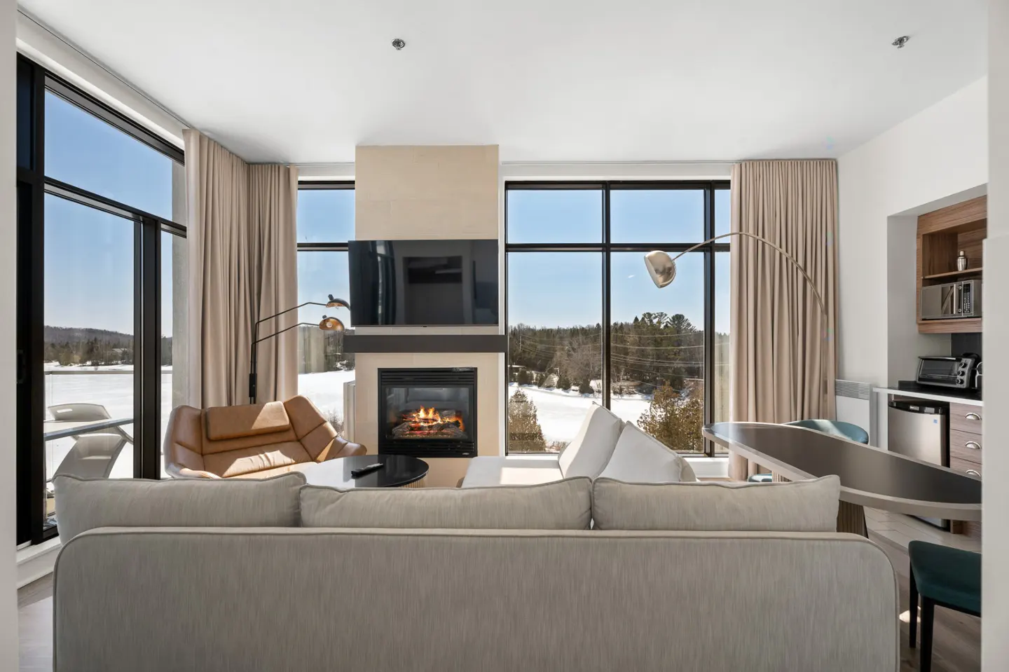 Bright living room with fireplace, TV, and large windows overlooking a snowy landscape. Beige sofa and curtains, with a kitchenette in the background.