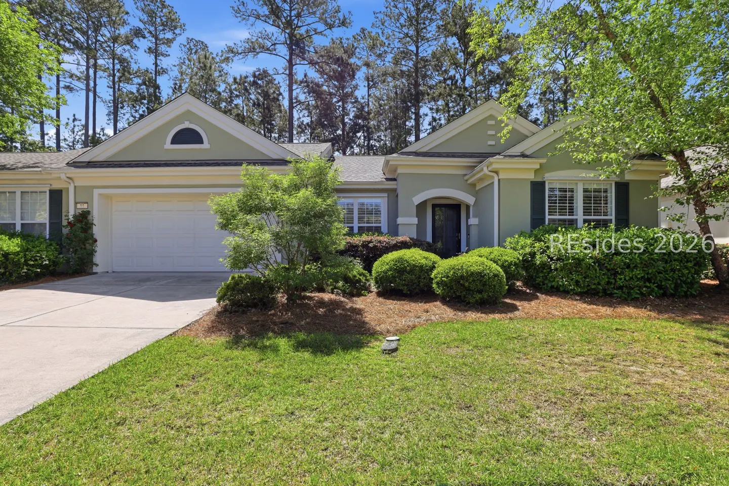 Exterior of a one-story, light green house with a white garage door, dark front door, and green lawn. Tall trees in the background.