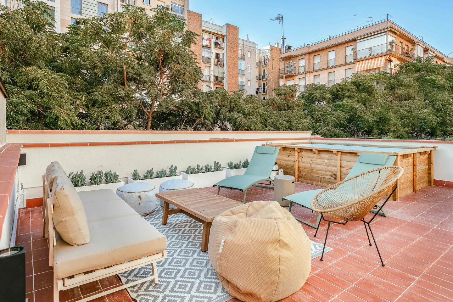 Rooftop patio with lounge seating, a wooden hot tub, and city views. The patio has a red tile floor and a gray and white patterned rug.