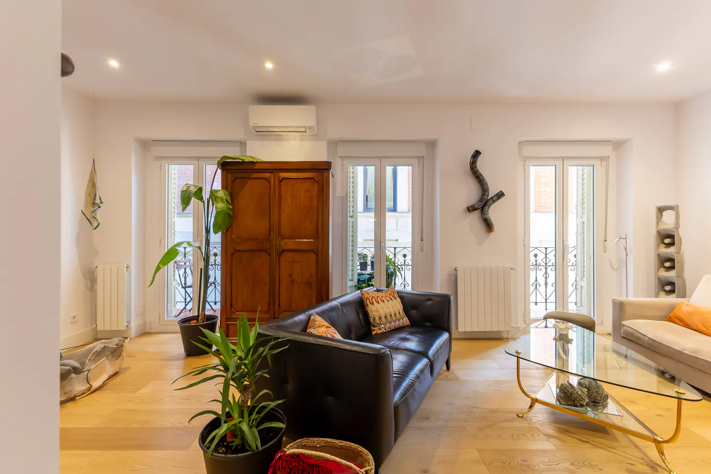 Bright living room with wood floors, white walls, and natural light. Features a black leather sofa, glass coffee table, and wooden wardrobe.