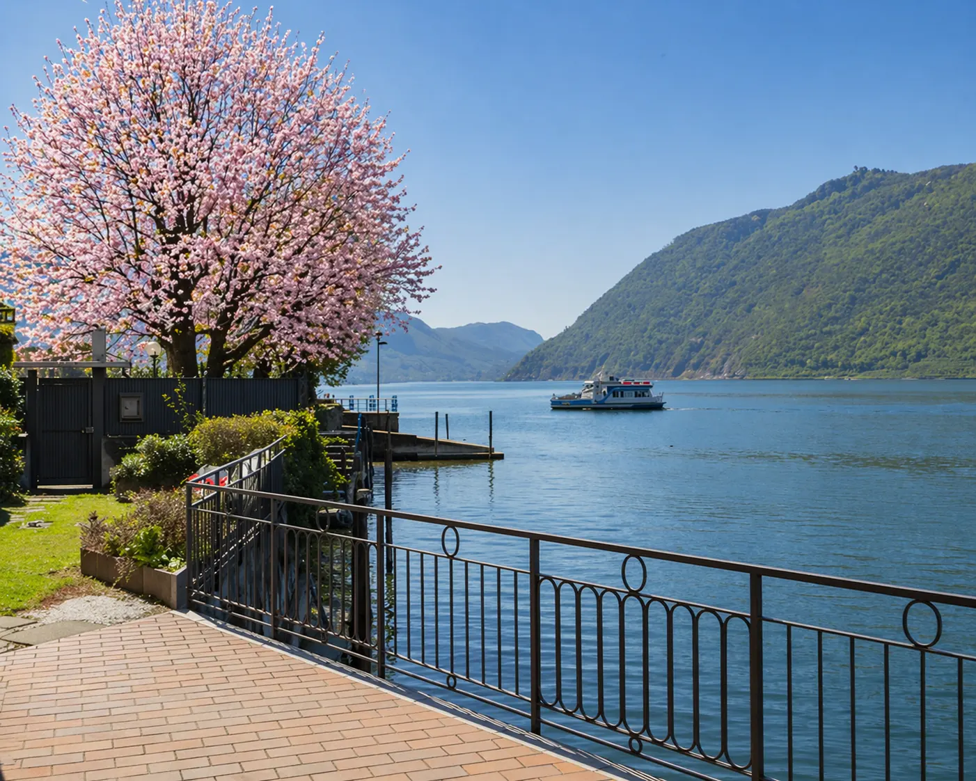 Waterfront view with a pink blossoming tree, blue water, mountains, and a boat under a clear blue sky.