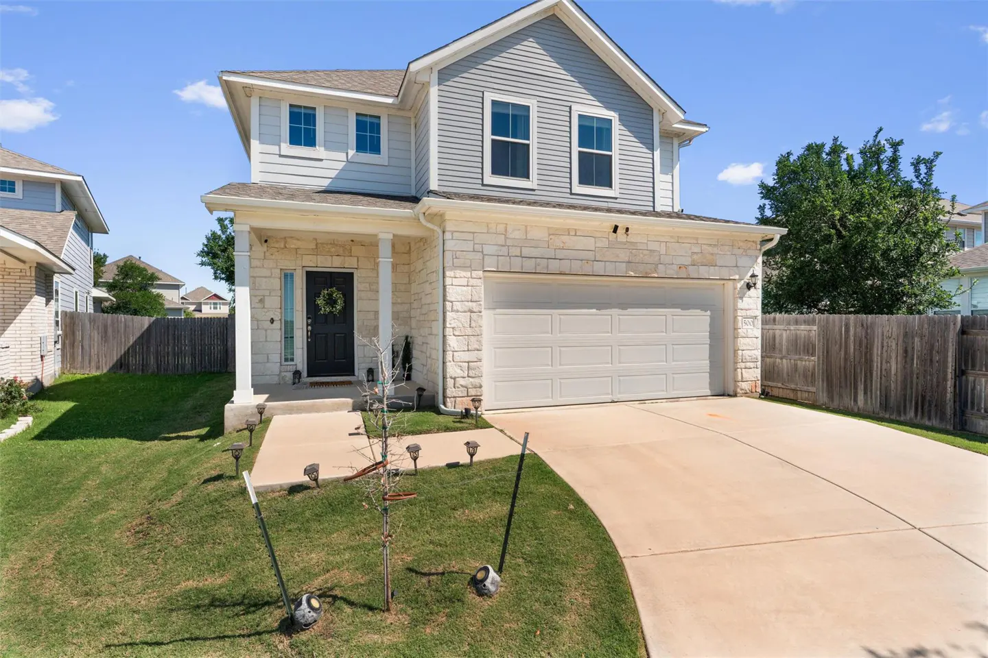 Two-story house with gray siding, stone facade, and white garage door. A black front door with a wreath is visible. Green lawn and driveway.