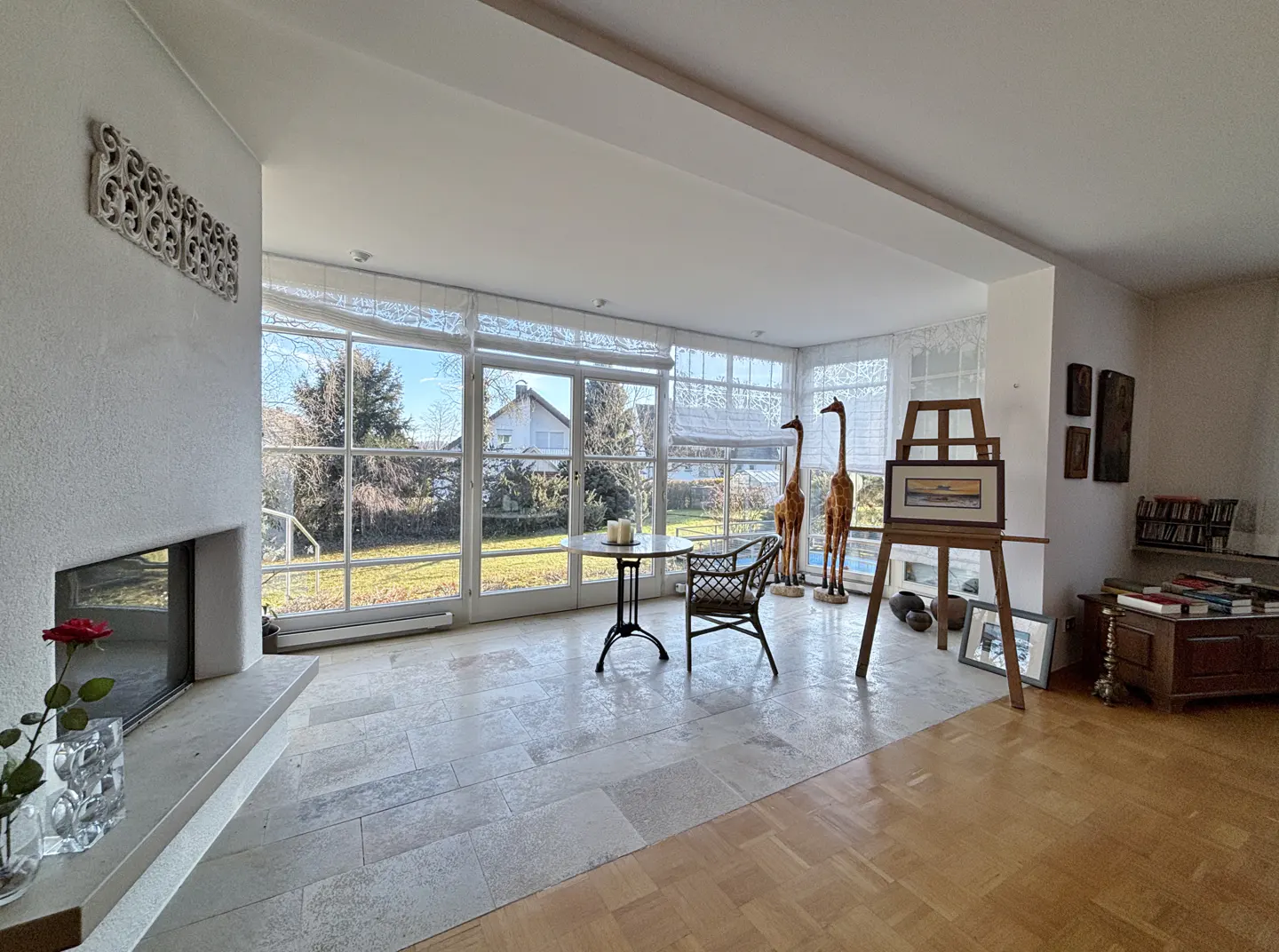 Bright sunroom with stone floors, large windows overlooking a green lawn, and interior decor including giraffe statues and an easel.