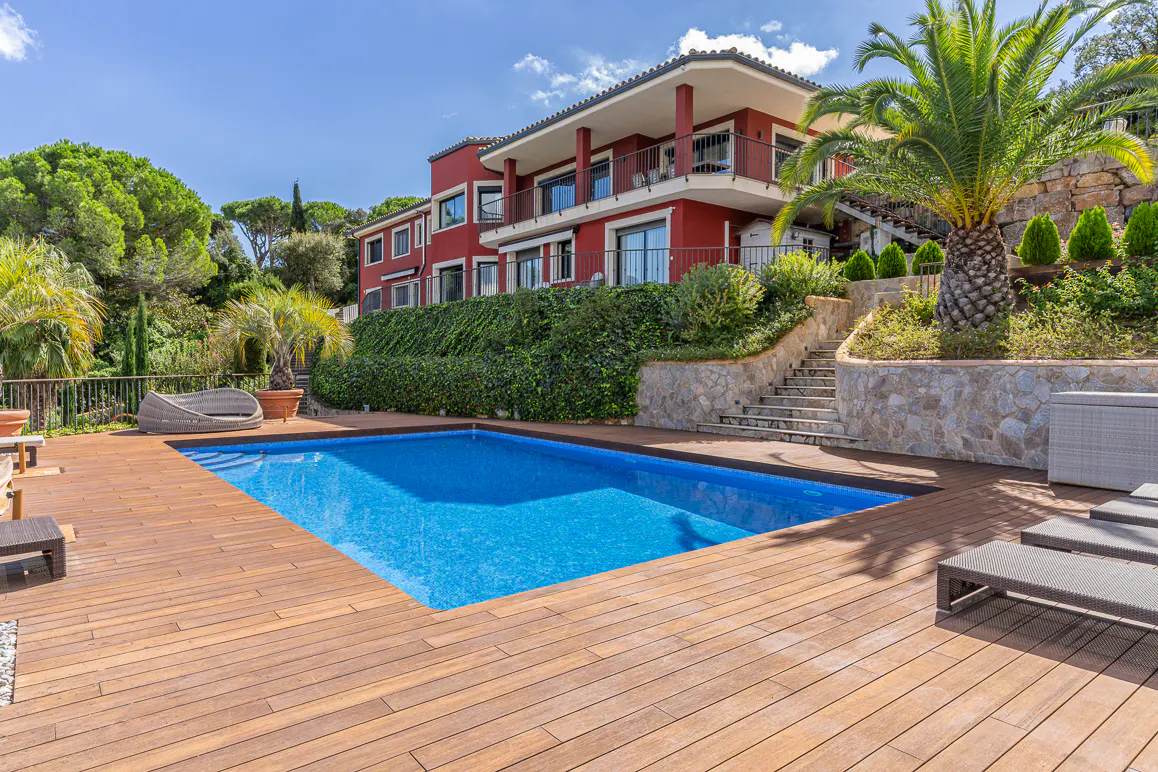 A red two-story house with a blue pool and wooden deck on a sunny day. Palm trees and greenery surround the property.