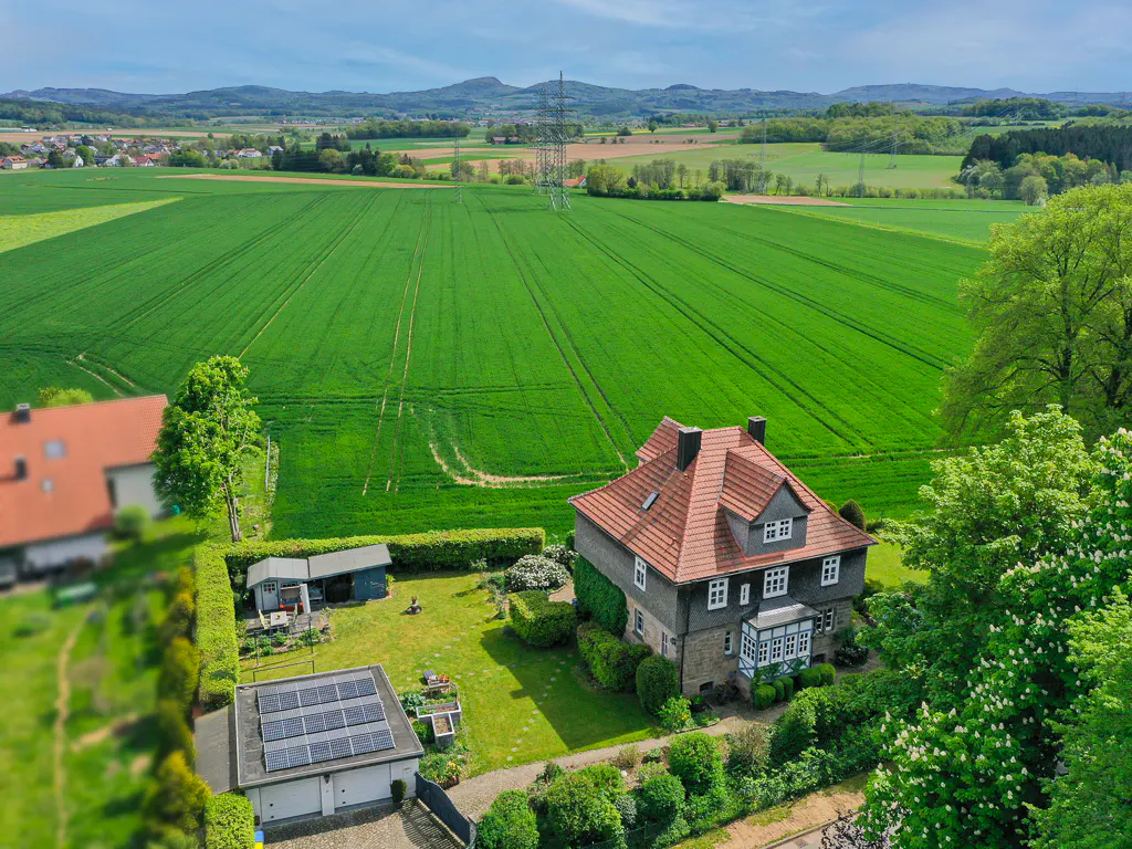 Aerial view of a house with a red tile roof, solar panels on a garage, and a large green field under a blue sky.