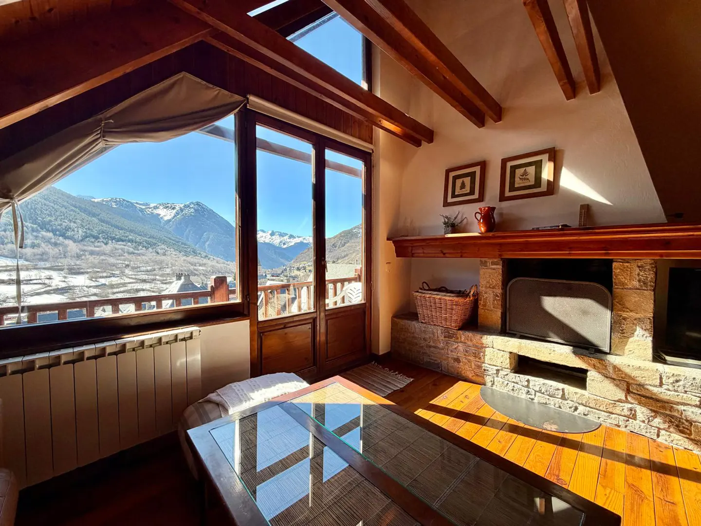 Attic living room with wood floors, exposed beams, and stone fireplace. Large windows offer a mountain view. A glass coffee table sits in the center.