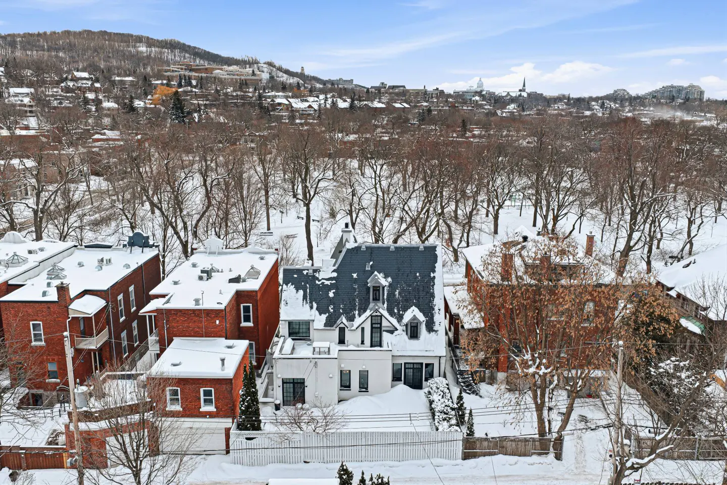 Aerial view of a white house with a dark roof, surrounded by snow-covered brick buildings and bare trees in winter.