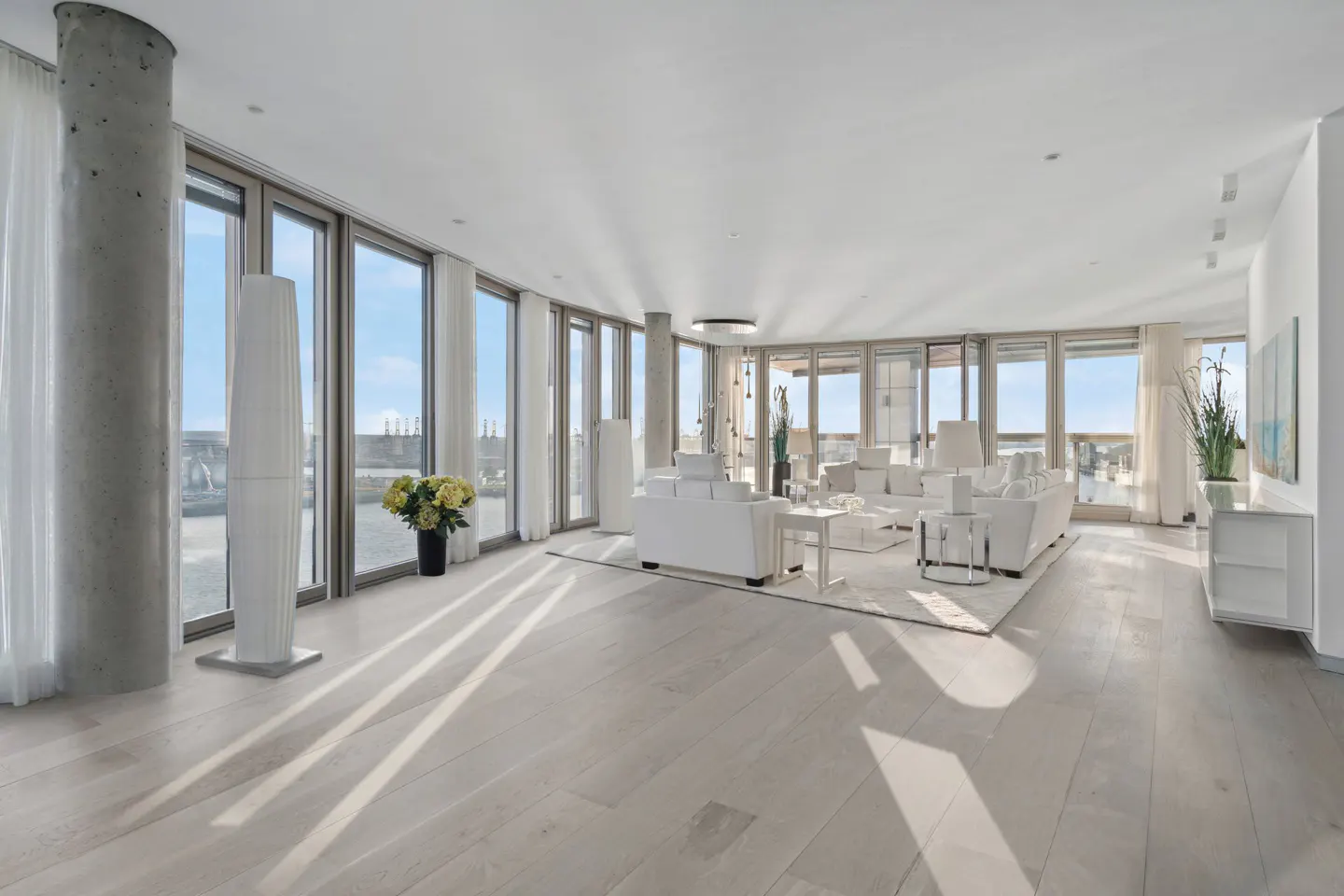Bright, modern living room with white furniture, light wood floors, and floor-to-ceiling windows offering a city view.