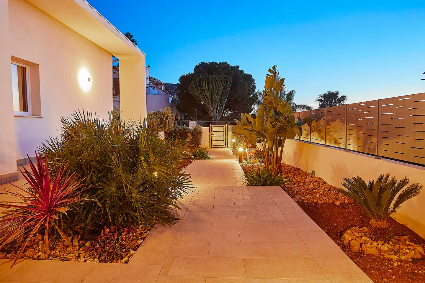 Exterior view of a modern home with a stone walkway, lush landscaping, and a wooden fence at dusk.