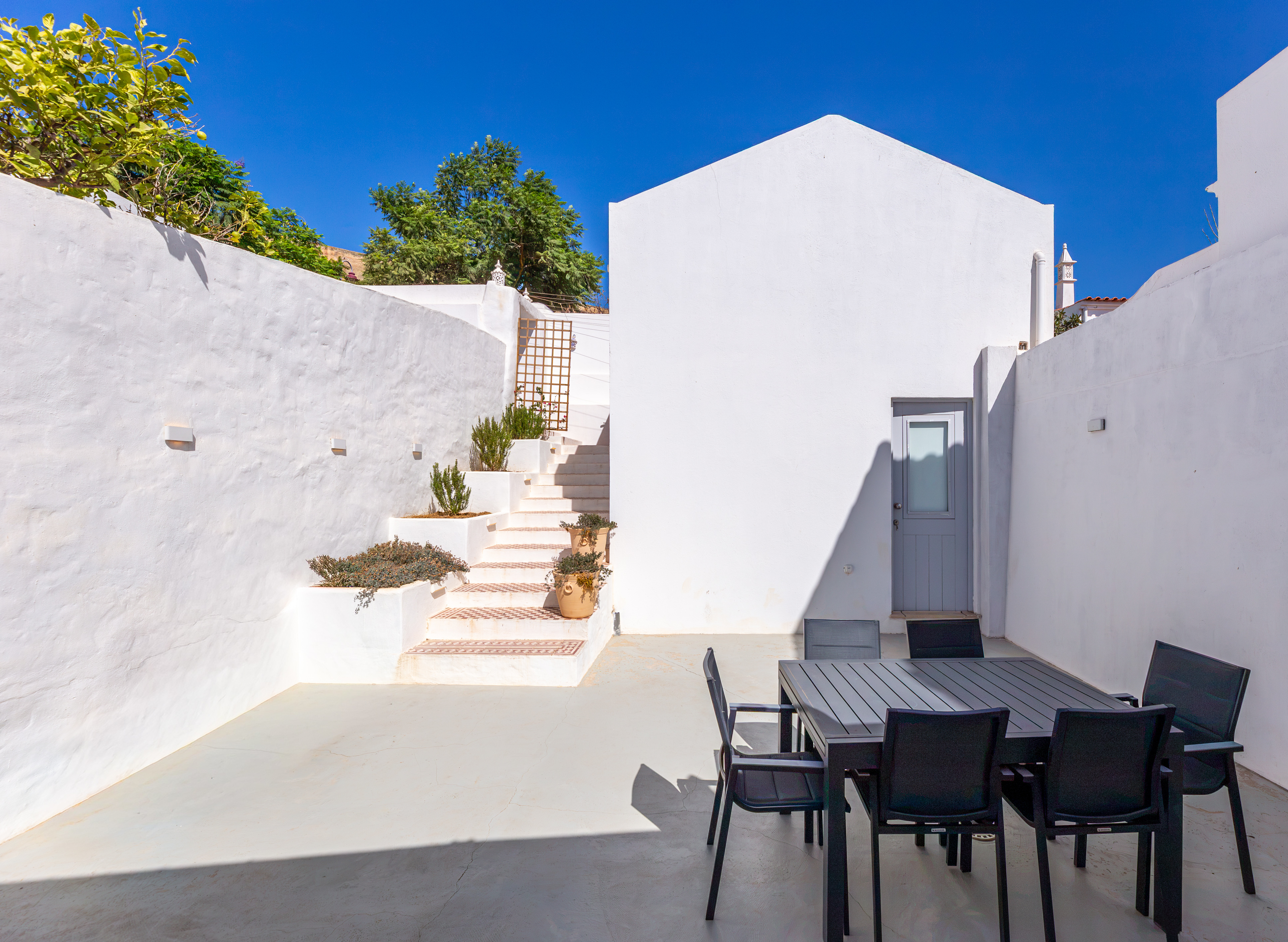 A white patio with a black dining set, stairs leading to a higher level, potted plants, and a clear blue sky. [AI-generated text]