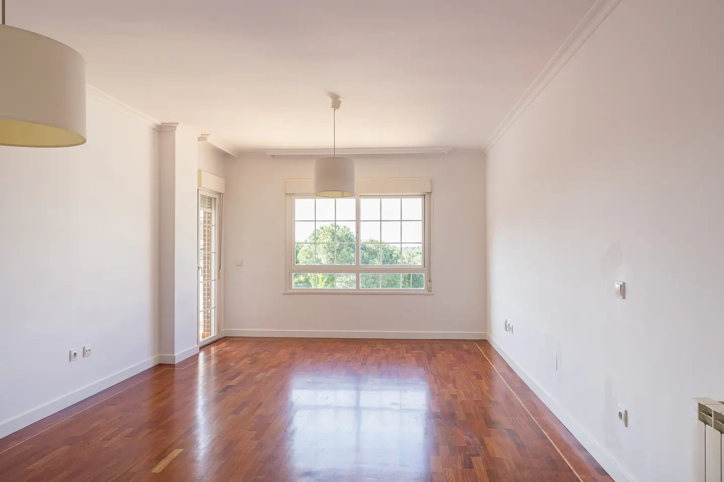 Bright, empty room with hardwood floors, white walls, and a large window overlooking trees. A white drum pendant light hangs from the ceiling.
