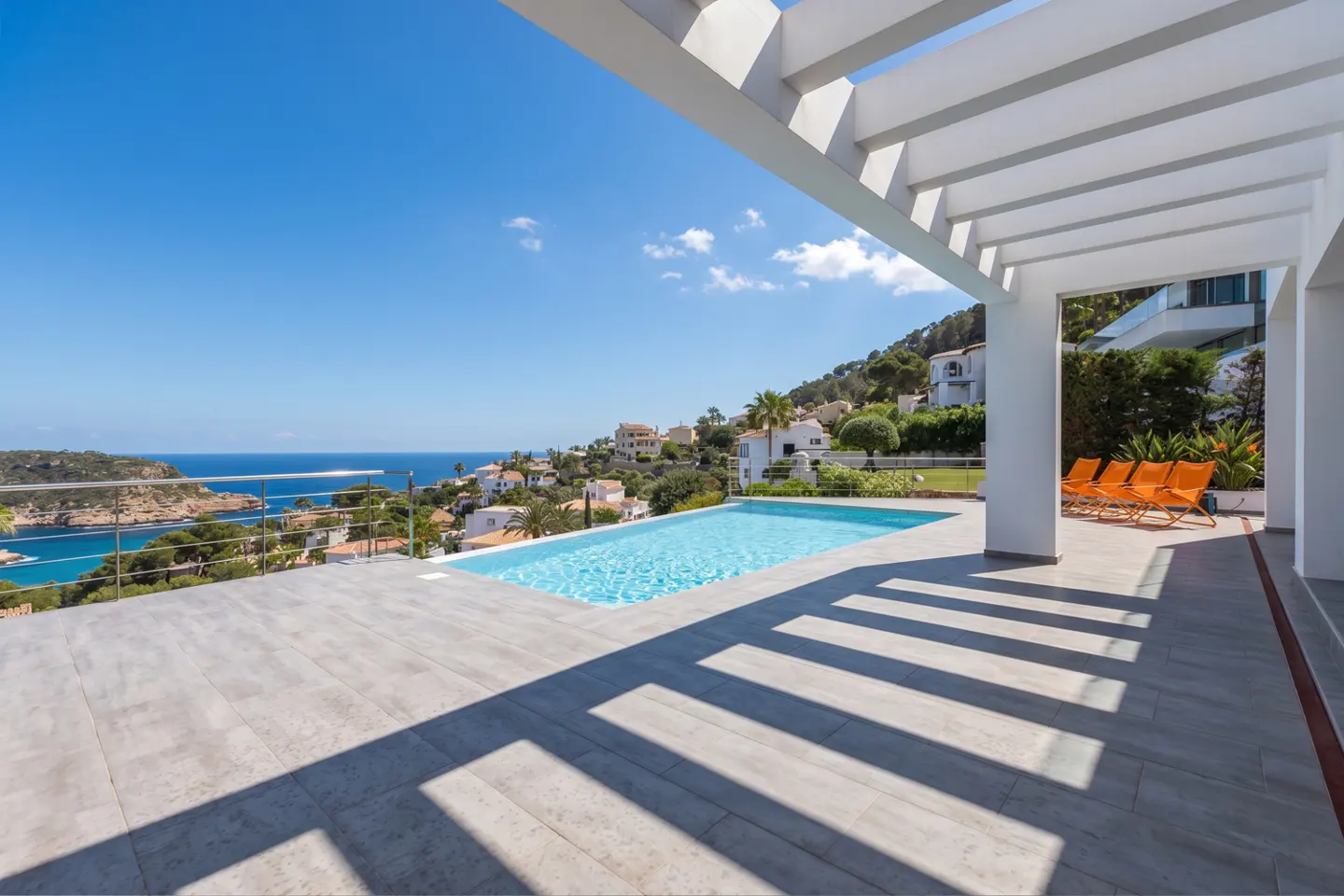 Luxury villa with pool overlooking the ocean. White pergola casts shadows on the gray tile patio. Four orange chairs are visible.