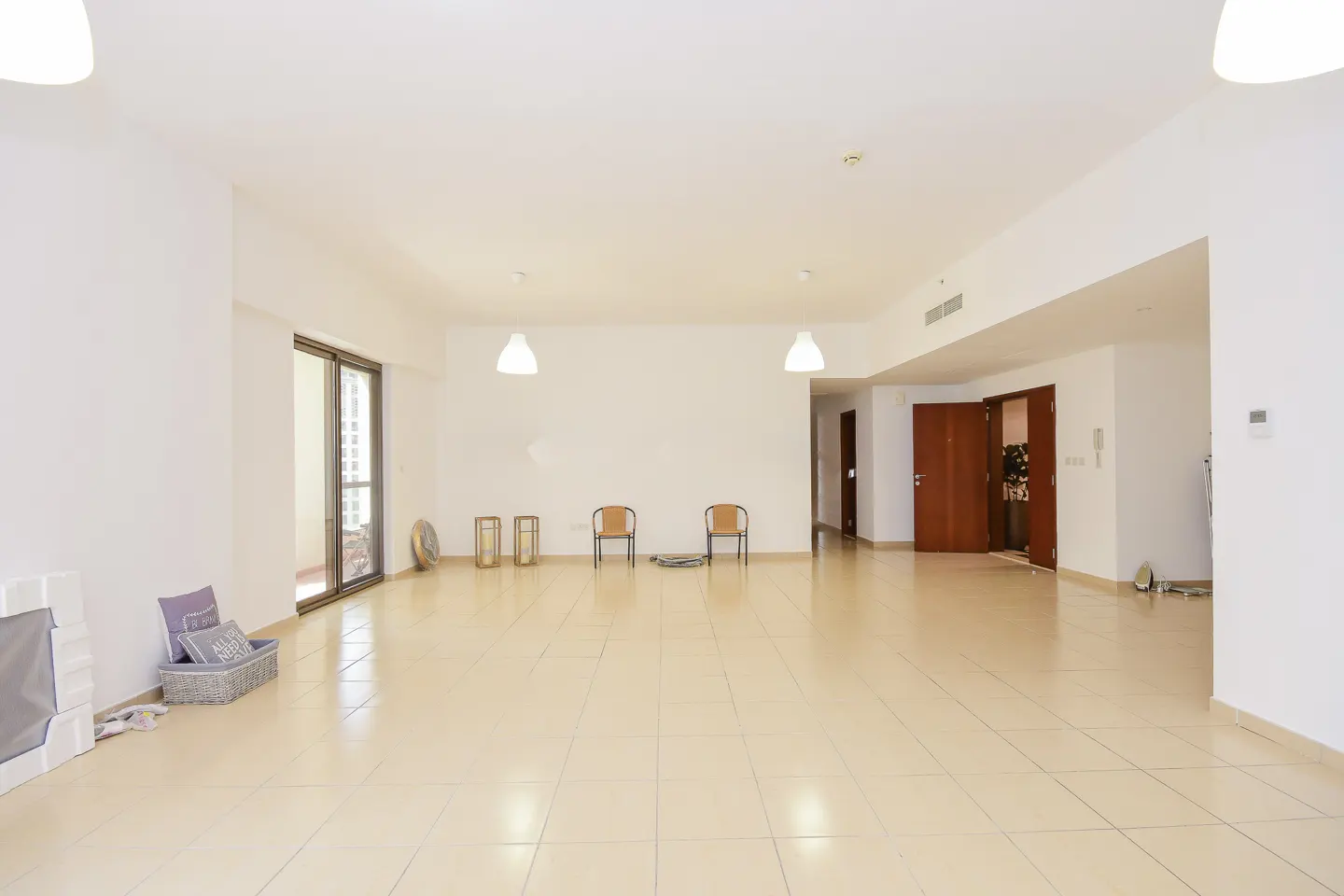 Bright, empty room with beige tile floor, white walls, and two chairs. Sliding glass doors lead to a balcony. Two brown doors are on the right.