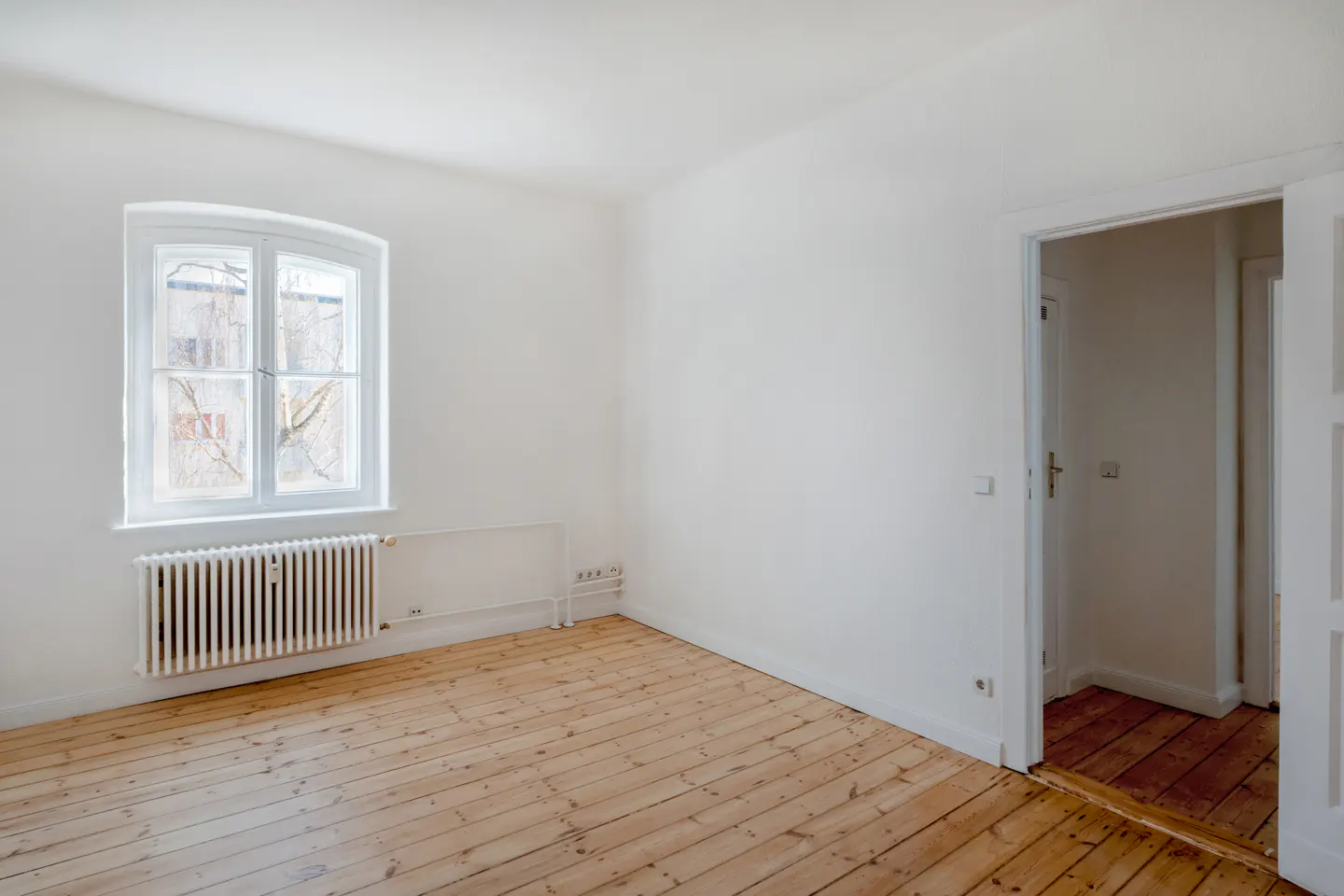 Bright, empty room with hardwood floors, white walls, and a window. A radiator sits beneath the window. An open doorway leads to another room.