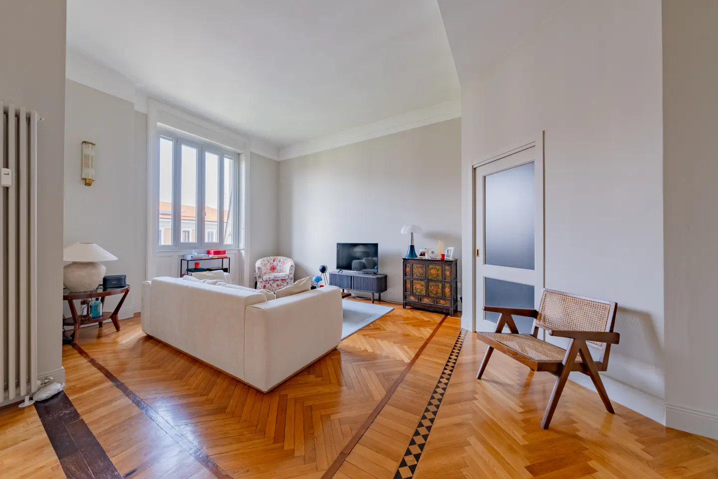 Bright living room with herringbone wood floors, white sofa, TV, and a wooden chair. A large window lets in natural light.