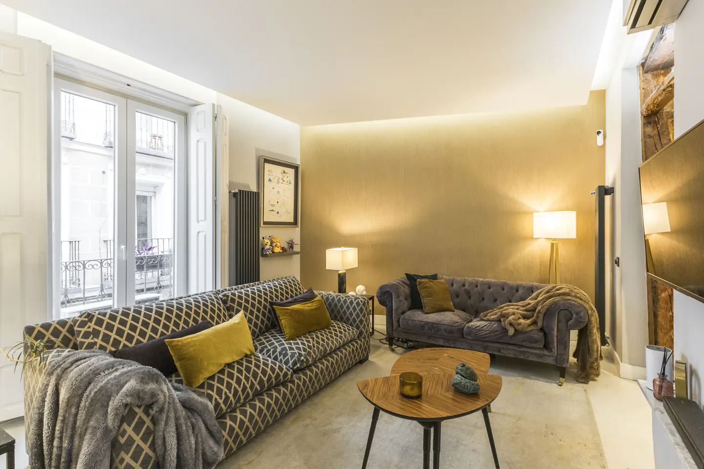 Living room with patterned sofa, grey velvet sofa, and wooden coffee table. Natural light from a window with white shutters.