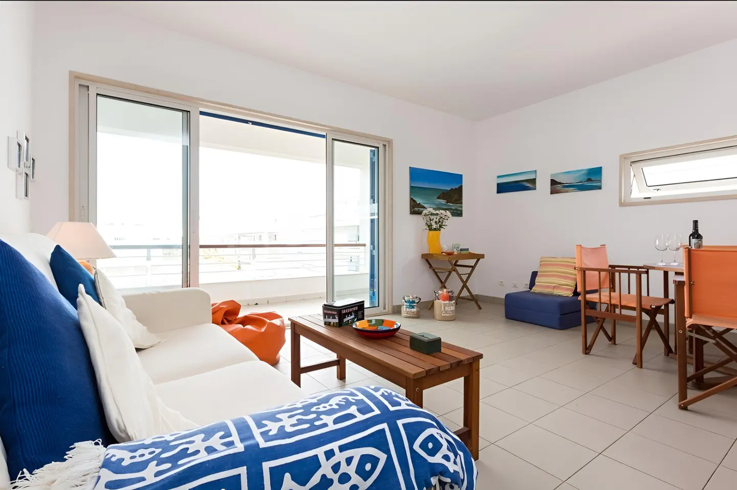 Bright living room with white walls, tile floor, and balcony. White sofa with blue pillows and blanket. Wooden table, chairs, and beach-themed art.