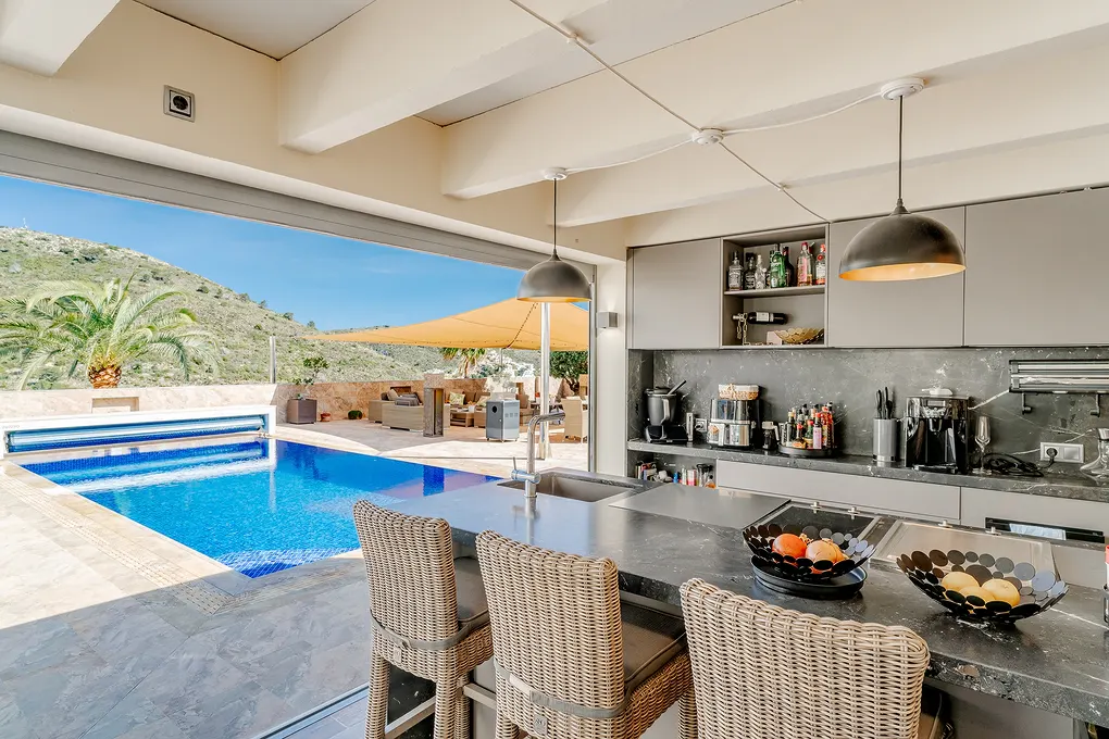 Open-concept kitchen with bar stools, gray cabinets, and black countertops. A pool and outdoor seating area are visible through a large sliding glass door.