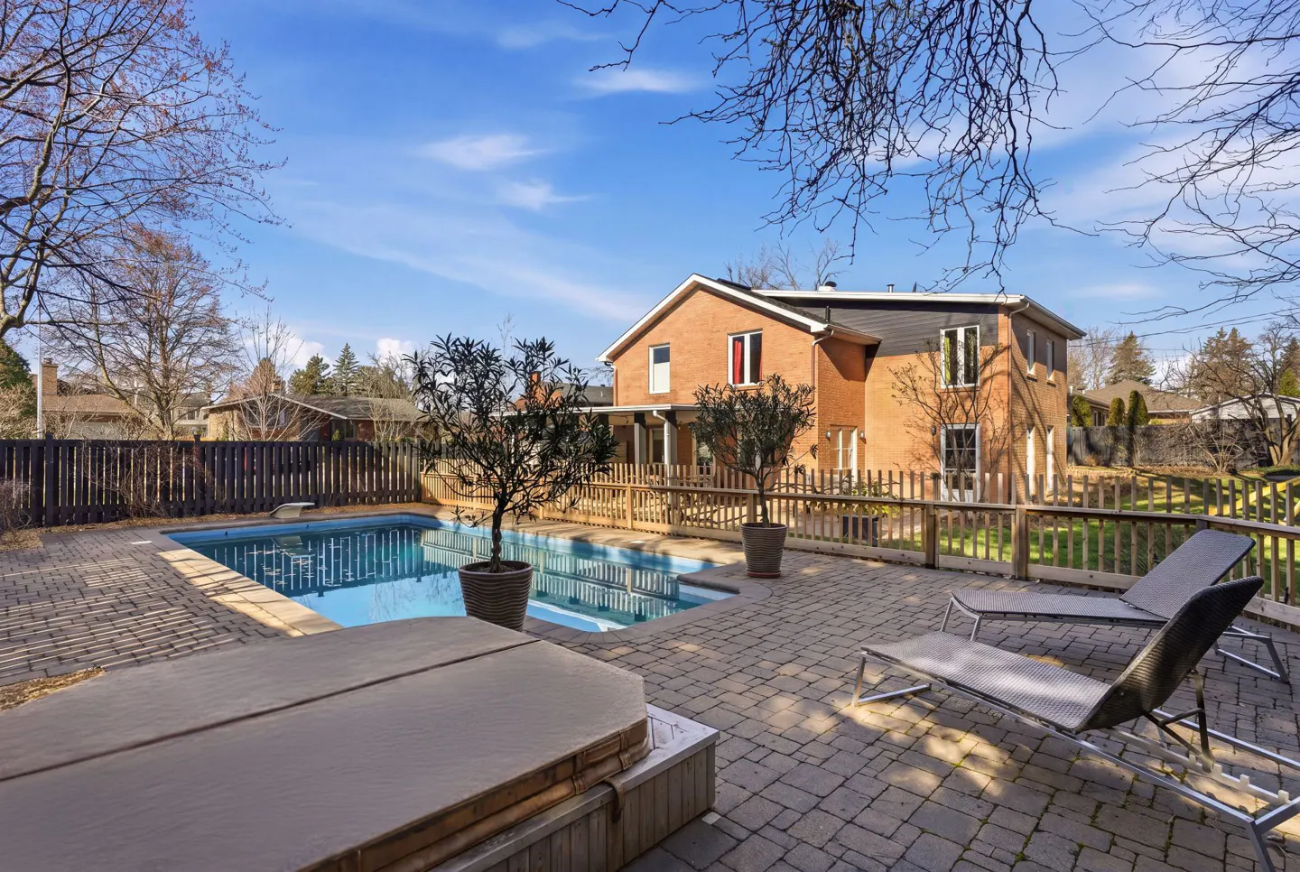 Backyard view of a brick house with a pool, patio, lounge chairs, and trees under a blue sky.