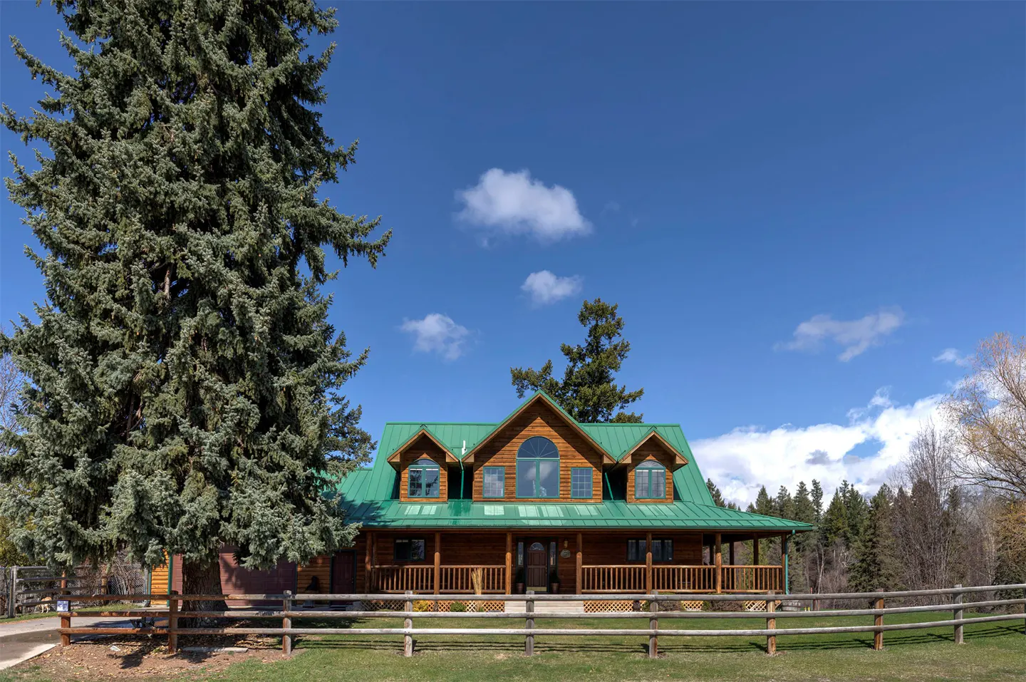 A log cabin with a green metal roof and a wooden porch sits behind a split rail fence on a sunny day. A large evergreen tree is to the left.