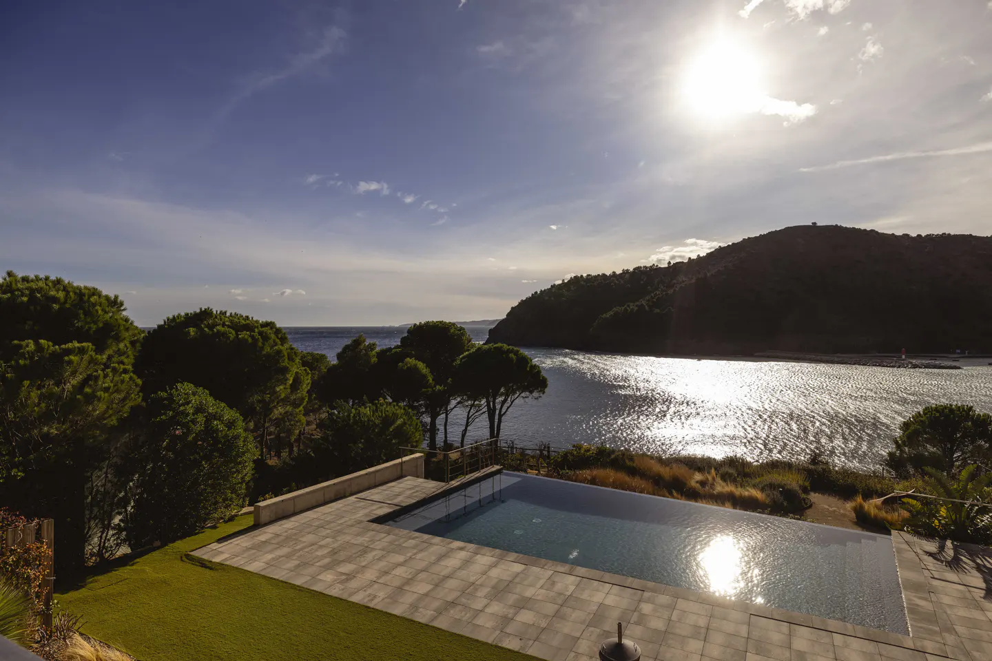 Scenic view of a pool overlooking a bay. The sun reflects on the water, with trees and a hill in the background.
