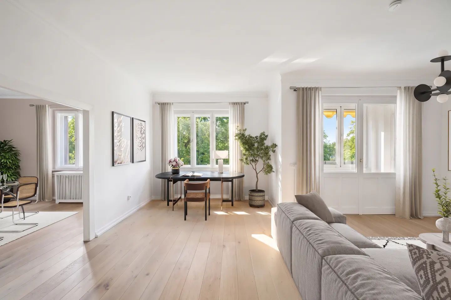 Bright, open living space with light wood floors, white walls, and large windows. A black desk and gray sofa add contrast.