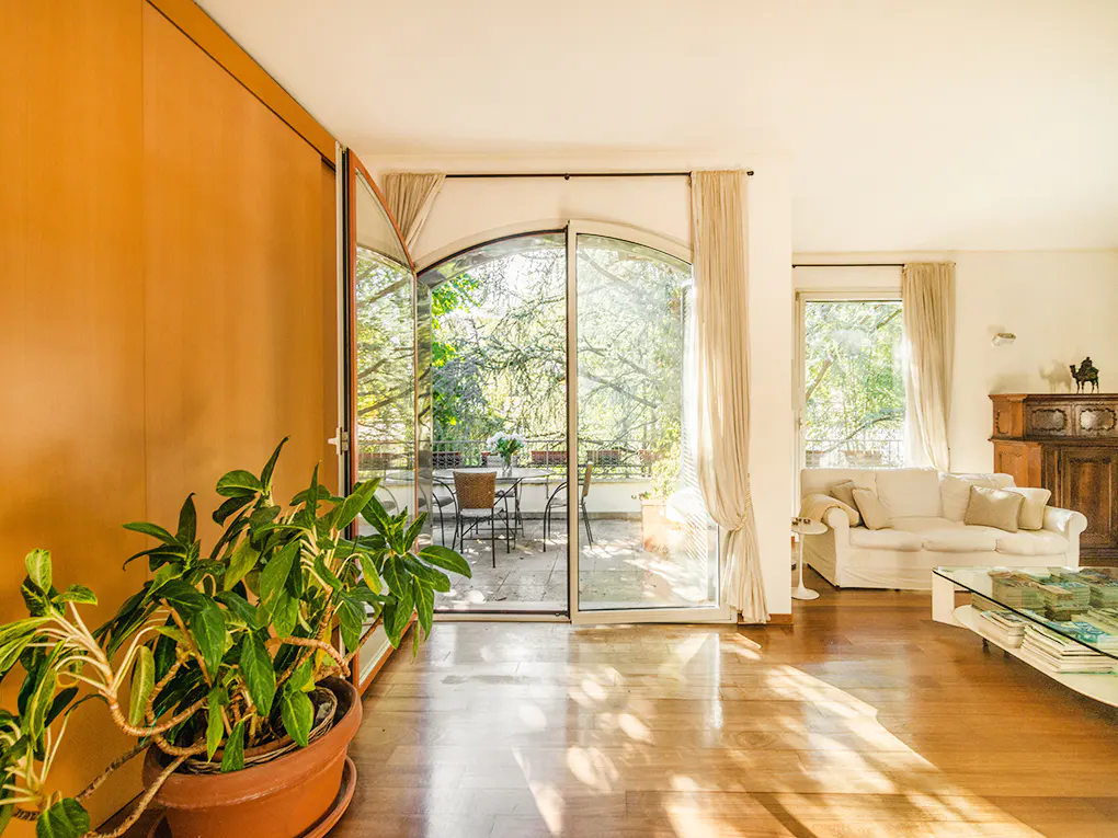 Bright living room with hardwood floors, a white sofa, and a glass coffee table. Sliding glass doors open to a balcony with a table and chairs. A potted plant sits in the foreground.