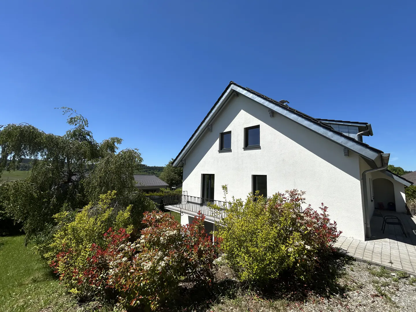 A white two-story house with a balcony, surrounded by green and red bushes under a clear blue sky.