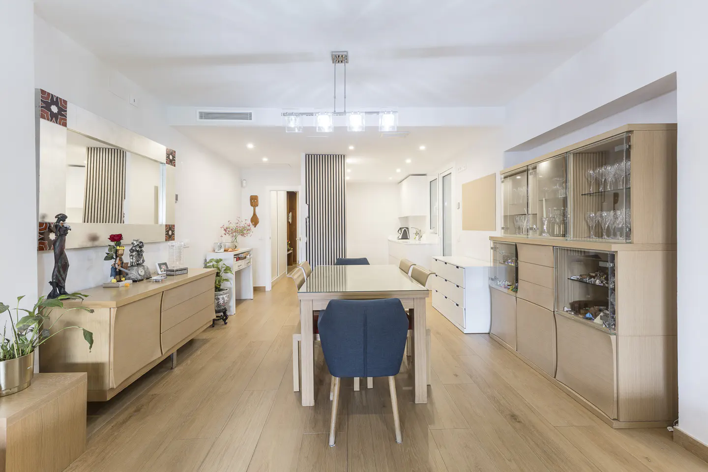 Bright dining room with light wood floors, a glass-top table, blue chairs, and a large wooden display cabinet.