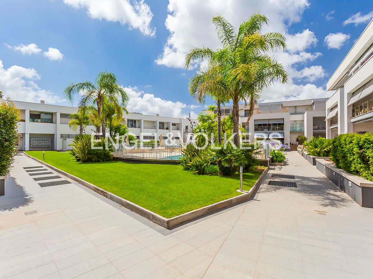 Modern apartment complex with a central green lawn, palm trees, and a swimming pool under a bright blue sky.