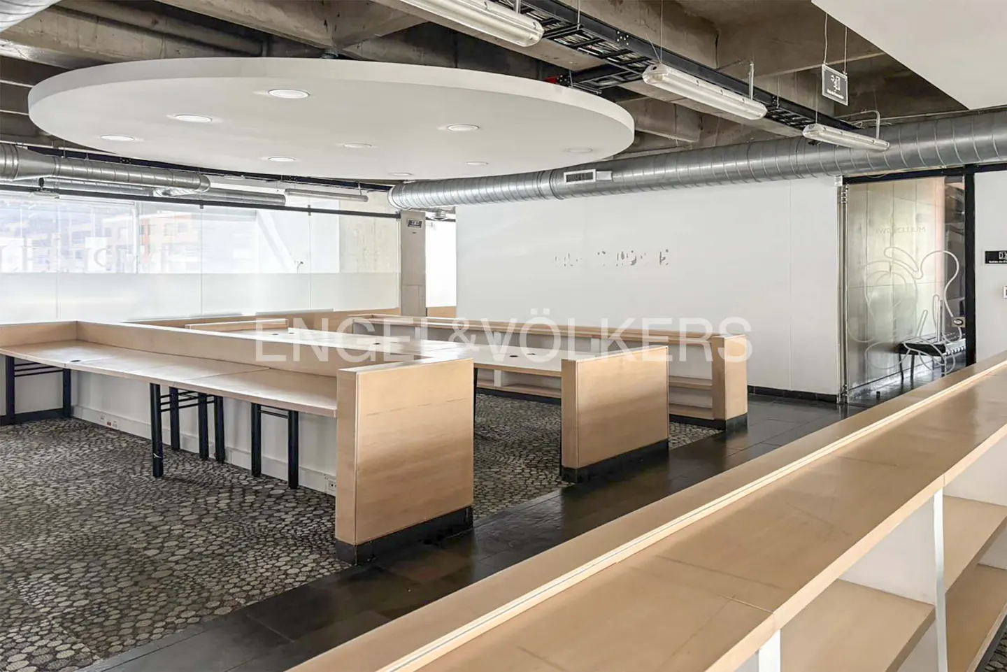 An open office space with light wood desks and black metal legs, a white wall, and a gray ceiling.