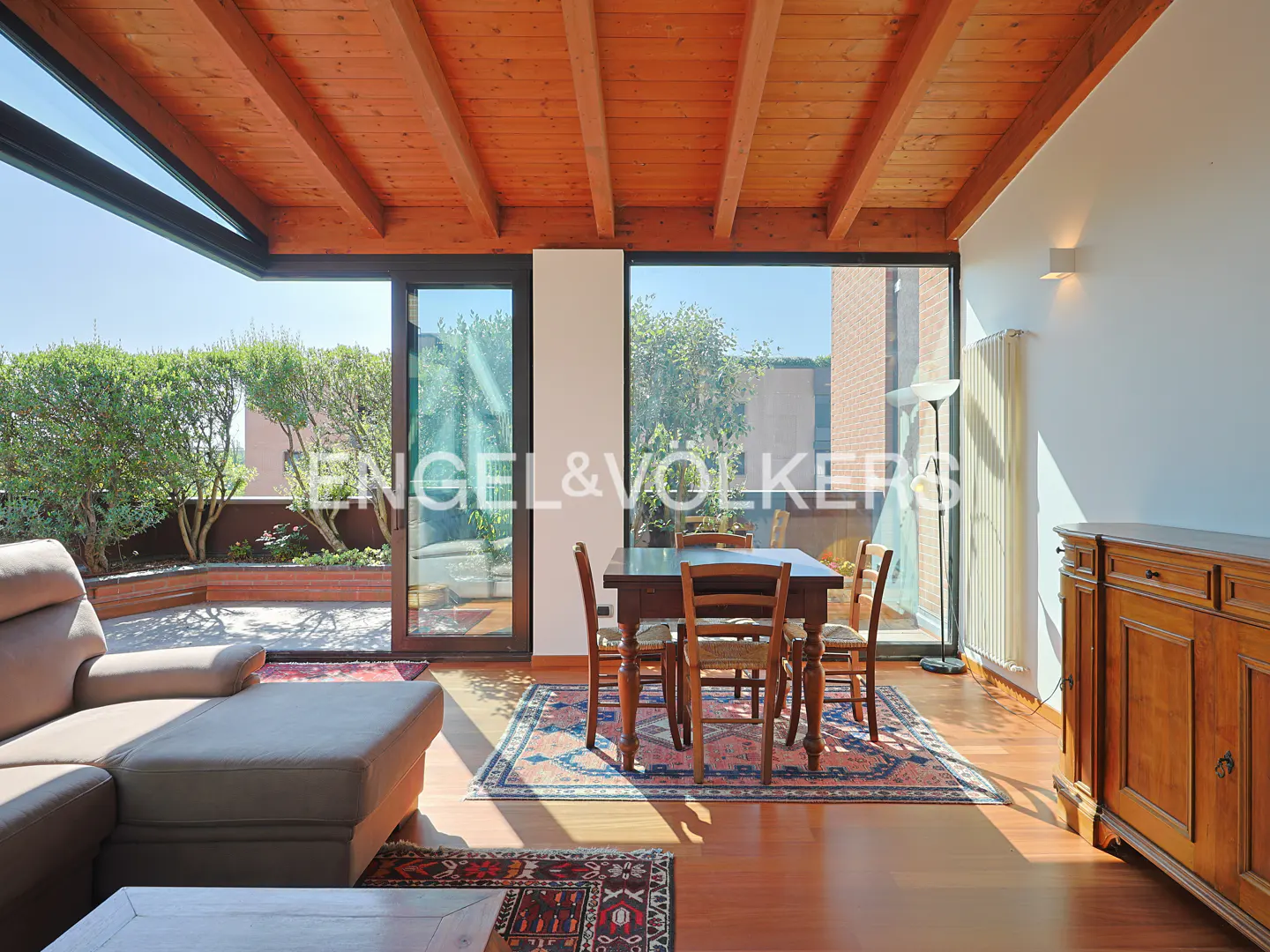 Living room with wooden ceiling, dining table, sofa, and large windows opening to a sunny terrace with greenery.