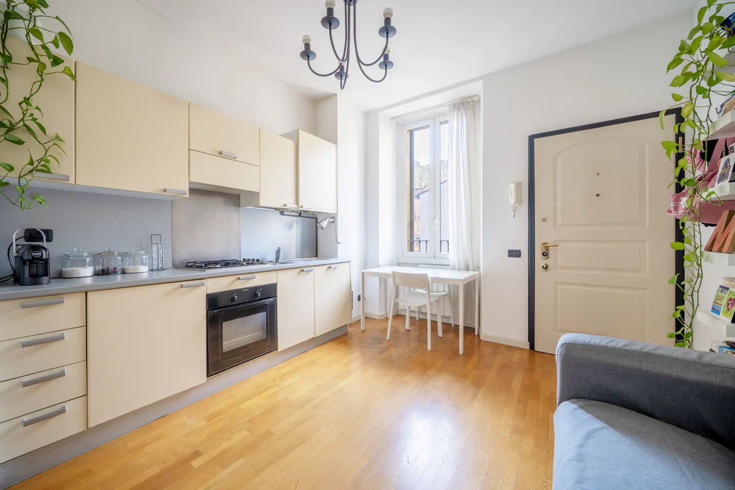 A bright kitchen with light wood floors, cream cabinets, and a black oven. A white table and chair sit by a window with sheer curtains. Green plants hang near the cabinets and door.