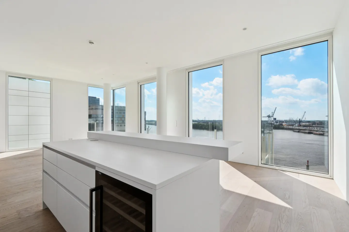 Bright, modern kitchen with white cabinets, island, and wood floors. Large windows offer a view of a harbor with cranes and blue sky.