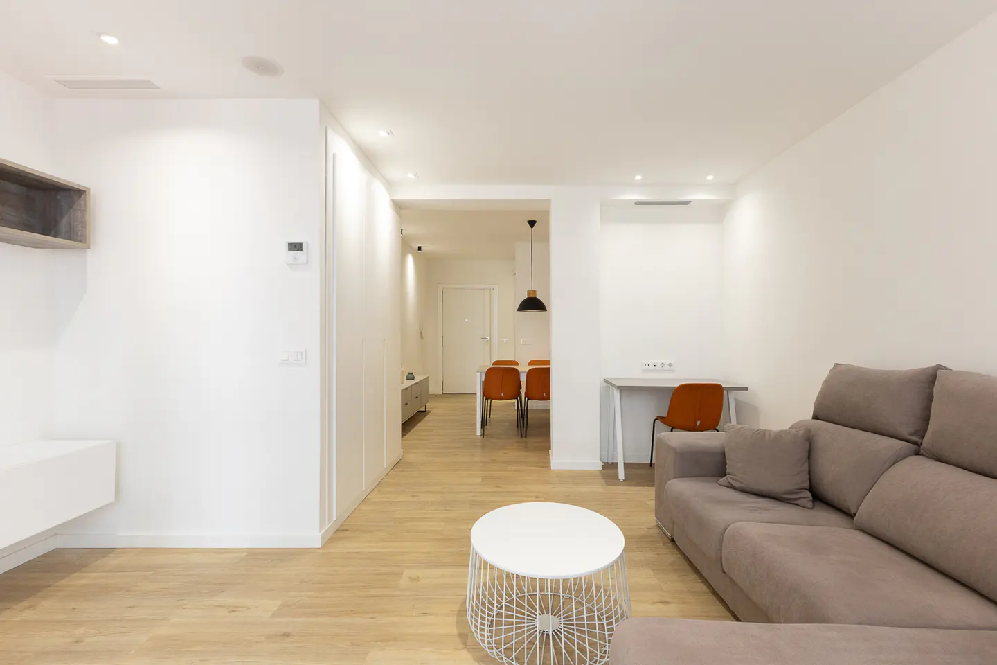 Modern living room with a grey sofa, white wire coffee table, and a view into a dining area with orange chairs.