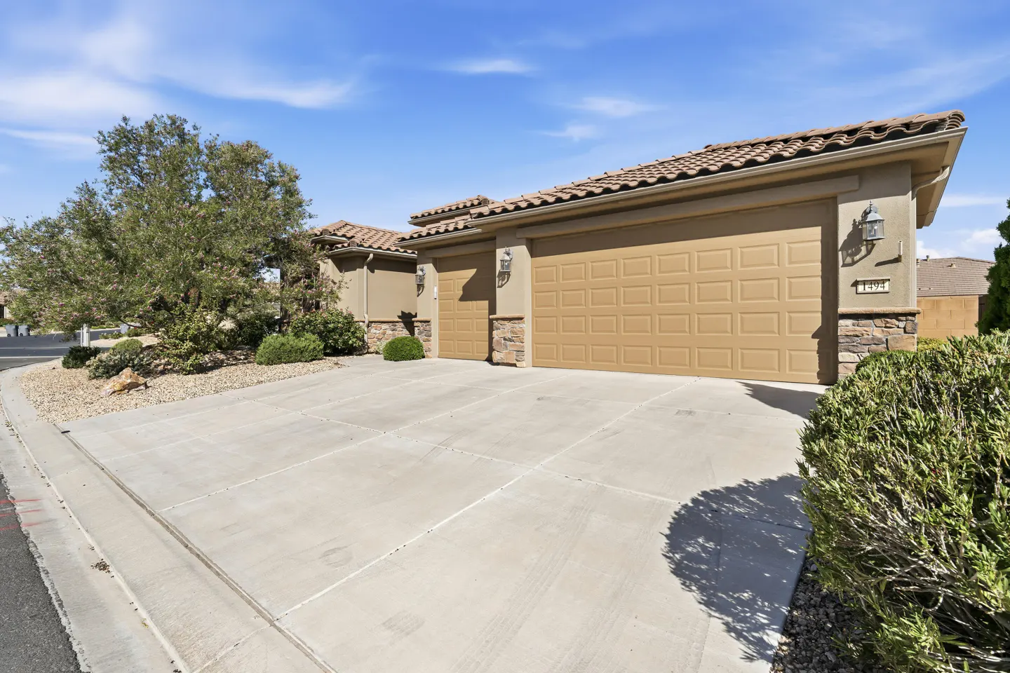 Tan stucco house exterior with a three-car garage, stone accents, tile roof, and a concrete driveway under a blue sky.