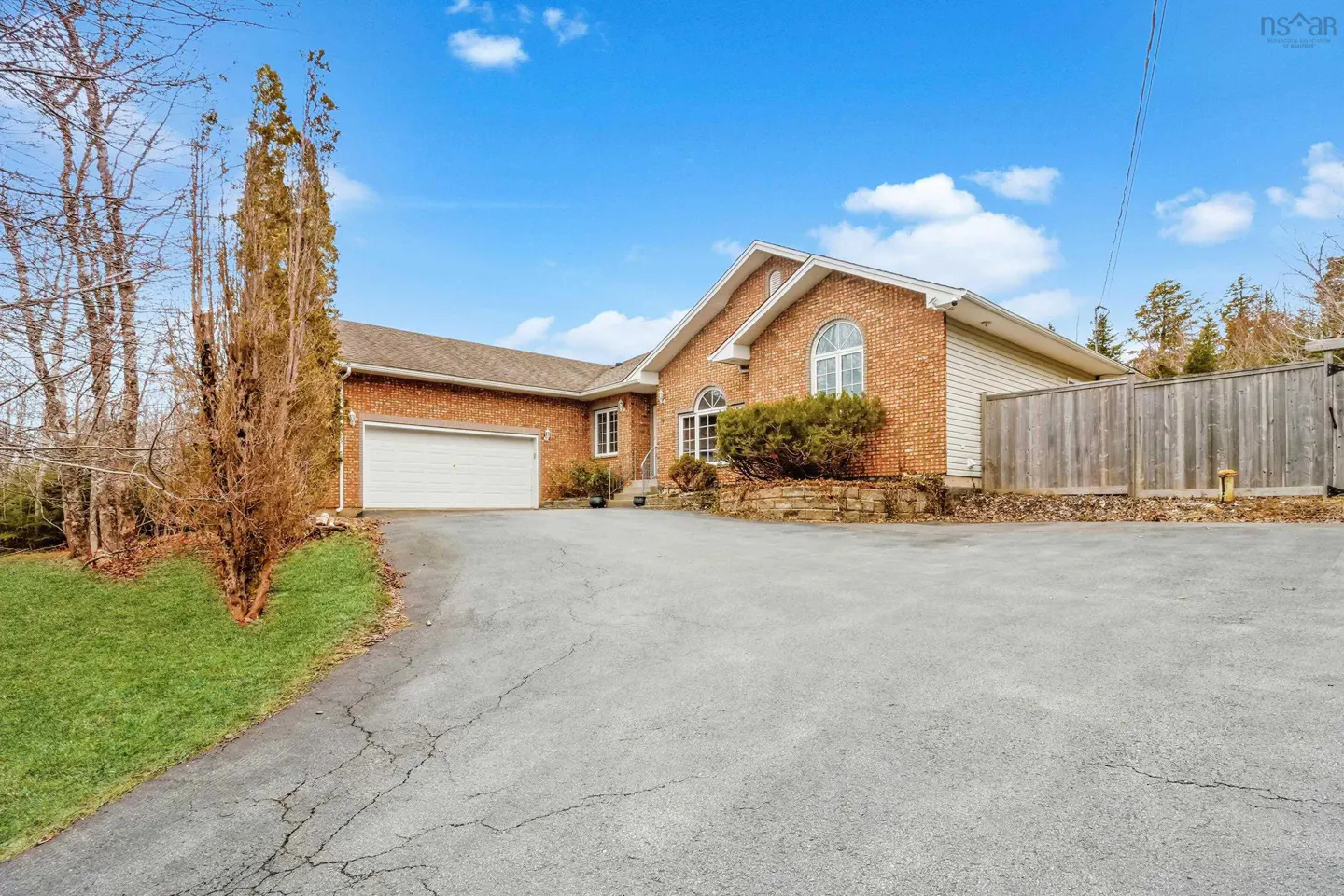 A single-story brick house with a white garage door and a large driveway under a blue sky.
