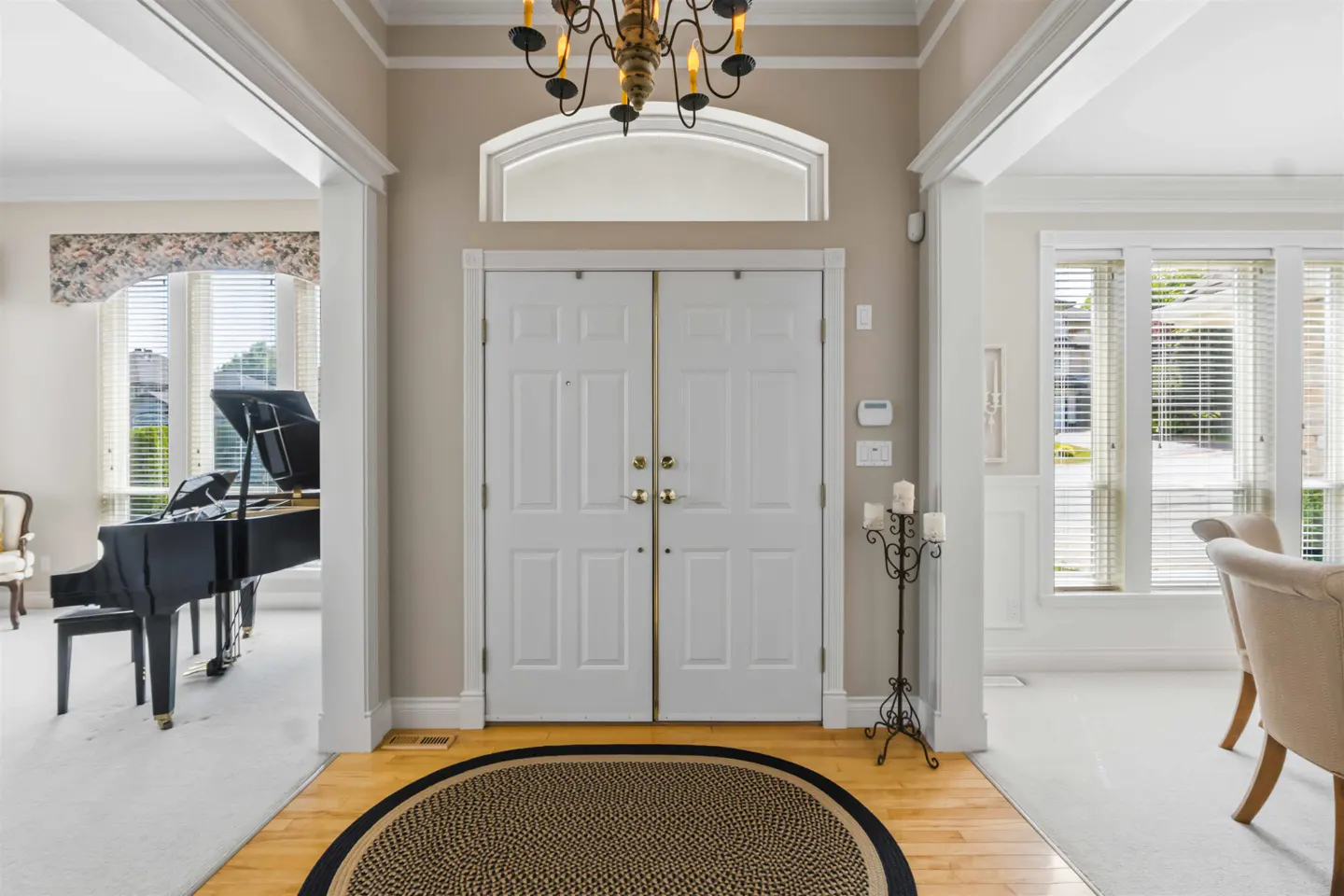 A foyer with double white doors, a gold chandelier, and a round rug on a wood floor. A piano and dining room are visible through archways.