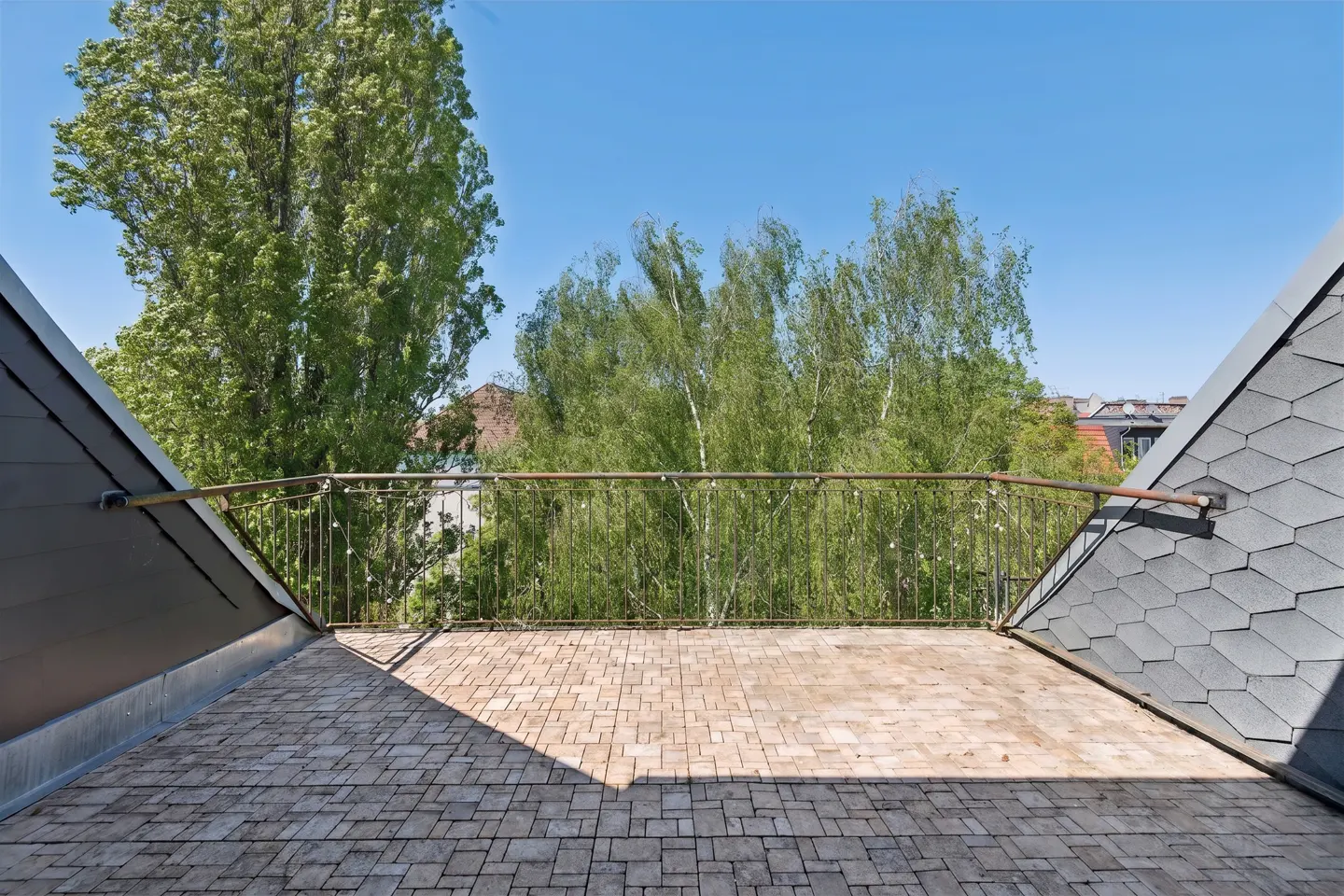 Rooftop terrace with wood-look tiles, metal railing, and gray shingled roof. Green trees and blue sky in the background.