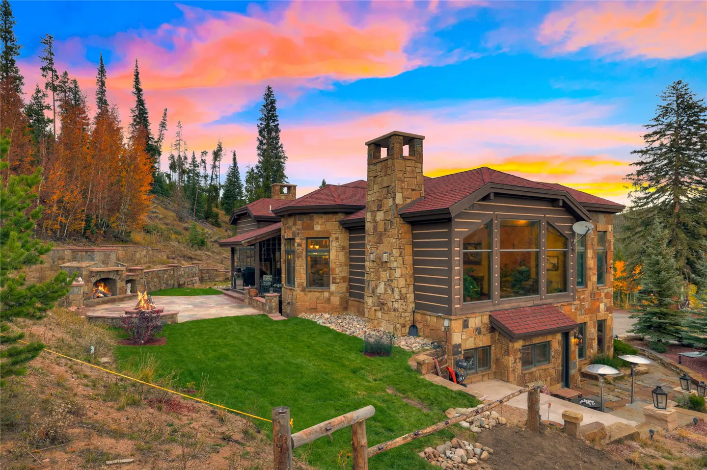 Exterior view of a stone and wood home with a red roof, fireplace, and colorful sunset.