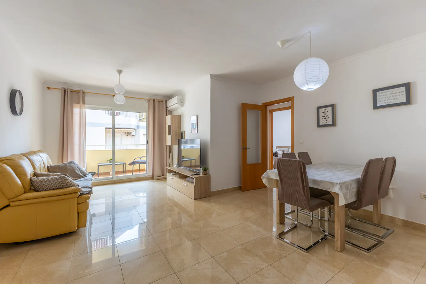 Bright living room with a yellow leather sofa, dining table with four chairs, and a balcony with a view.