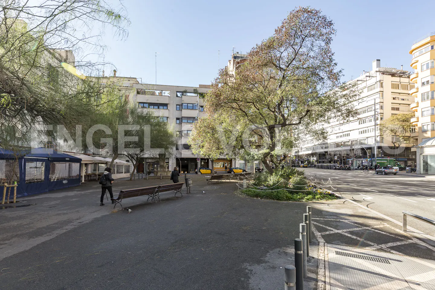 City square with benches, trees, and buildings. People walk by shops with blue awnings. Yellow taxis are parked in the background.